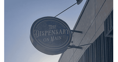 Street view of The Dispensary on Main storefront in downtown Galesburg, Illinois, with the shop’s hanging sign visible against the morning sun.